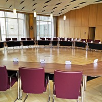 Bild vergrößern: Ein Blick in den Sitzungssaal. Zu sehen sind Sthle und Tische, die in einem groen Kreis vor einer groe Fensterfront aufgestellt sind.