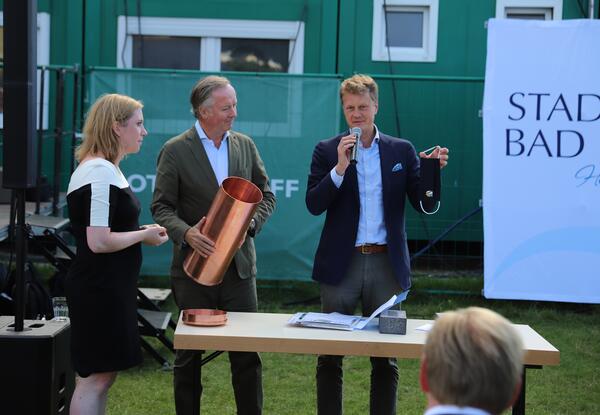 Auf dem Bild stehen Frau Zweig, Herr Wulff und Dr. Brinkmann an einem Tisch. Herr Wulff hält die Zeitkapsel in den Händen. Der Bürgermeister erläutert den Inhalt und hält einen Mund-Nasen-Schutz hoch.